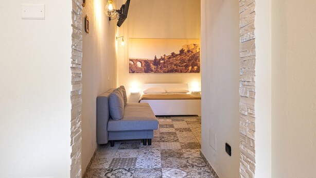 Um corredor estreito com parede de pedra, um sofá cinzento, uma cama com roupa de cama branca e um quadro grande emoldurado na parede.