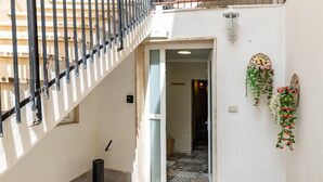 Uma escada com guarda-corpo de metal, uma porta que dá para um espaço interno e uma luminária fixada na parede.