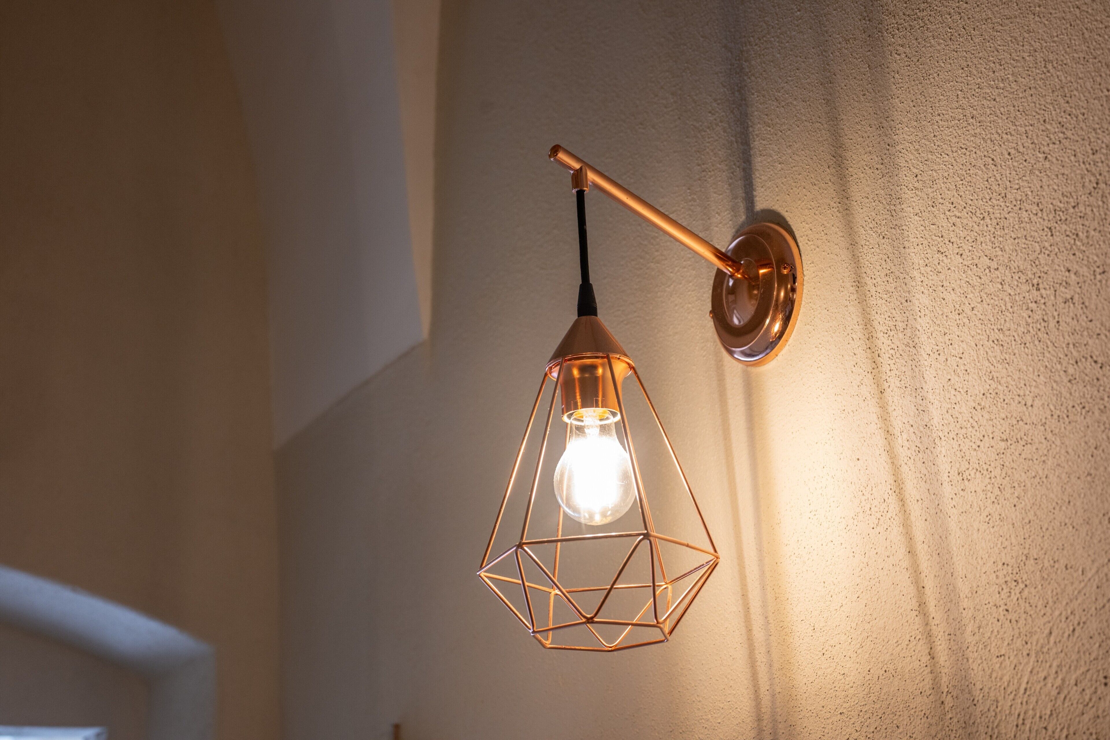 Lampada a sospensione geometrica con lampadina a vista, fissata a una parete dalla superficie strutturata.