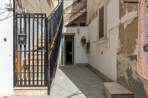 Une ruelle étroite, avec un portail en métal noir, un mur blanc dont la peinture s’écaillait et une petite fenêtre agrémentée d’un arrangement floral.