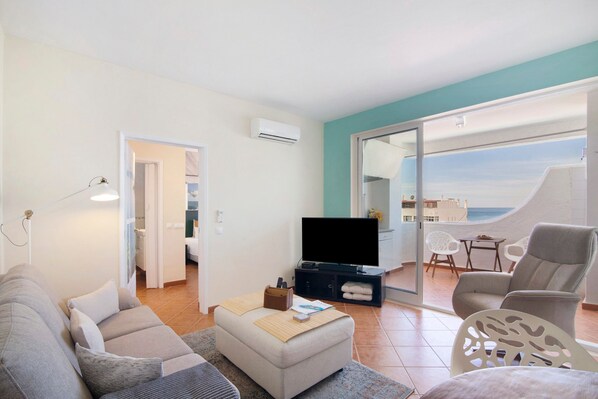 A living room with a sofa, ottoman, flat-screen TV, and a view of the sea through a sliding glass door.