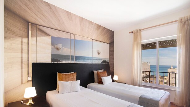 Une chambre d’hôtel avec deux lits, une table de chevet avec une lampe, une grande fenêtre donnant sur la mer et une œuvre d’art accrochée au mur.
