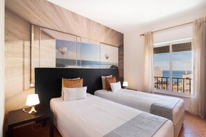 A hotel room with two beds, a nightstand with a lamp, a large window with a view of the sea, and a wall-mounted artwork.