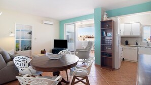 Un salon comprenant une table à manger, des chaises blanches, un canapé, un téléviseur et un coin cuisine avec des placards et un évier.