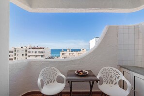 A balcony with two white chairs, a small table with a plate of food, and a view of the sea and buildings.