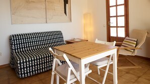 Een kamer met een houten tafel, witte stoelen, een gestreept bankje, een groot abstract schilderij, een lamp en een raam met jaloezieën.