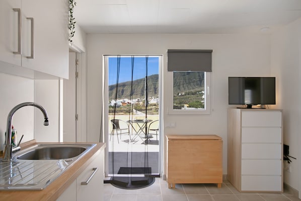 A modern kitchen with a sink, a wooden cabinet, a television on a stand, and a view of a mountainous landscape through a glass door.