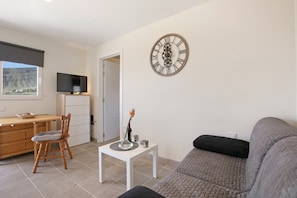 A living room with a sofa, a television on a stand, a dining table with chairs, and a decorative wall clock.