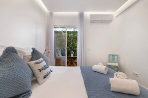 A modern hotel room with a bed, a small table, a chair, and a view of a balcony with plants.