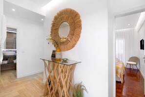 Un intérieur moderne avec une console en verre, un miroir tressé et un lit recouvert d’une couverture à rayures.
