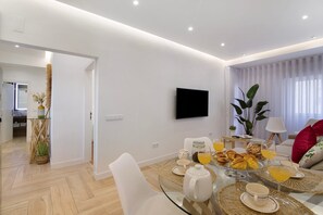 A modern living room with a glass dining table, white chairs, a flat-screen TV, and a sofa with a red cushion.
