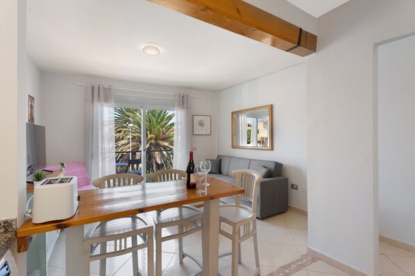 Uma área de refeições com mesa e cadeiras de madeira, um sofá, um pequeno espaço de cozinha com uma torradeira e uma varanda com vista para palmeiras.