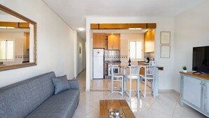 Un salon avec un canapé, une table basse et un téléviseur. Il y a également un coin cuisine avec des meubles blancs et une table à manger avec des chaises.