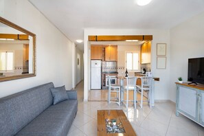 A living room with a sofa, a coffee table, and a TV. There is a kitchen area with white cabinets and a dining table with chairs.