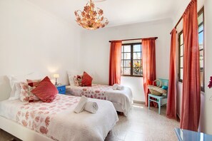Habitación de hotel con dos camas, una araña, una ventana con cortinas, una silla con una cesta y una mesita de noche con lámpara.
