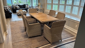 A dining area with a wooden table and wicker chairs, a patterned rug, and a view of the outside through large windows. - Sunny, Beach Days - Enjoy the beach just steps away in Clinton, Connecticut  (Clinton)