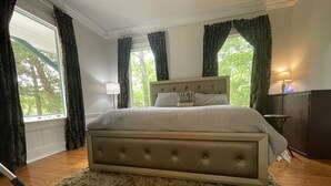 Un dormitorio con cama de cabecera capitonada, una alfombra mullida, una mesita de noche con lámpara y vistas a árboles por la ventana.