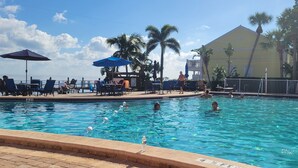 Ein Pool mit Liegestühlen und Sonnenschirmen, umgeben von Palmen und einem Gebäude im Hintergrund.