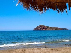 A beach with a rocky island in the distance and a thatched roof structure in the foreground.