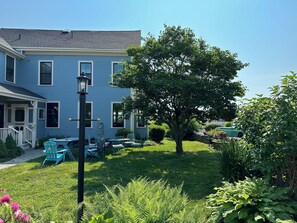 A two-story blue house with a porch, outdoor seating, and a large tree in the yard.