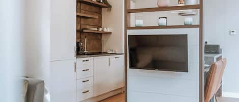 Una cocina moderna con armarios blancos, un televisor integrado y un estante de madera con objetos decorativos.