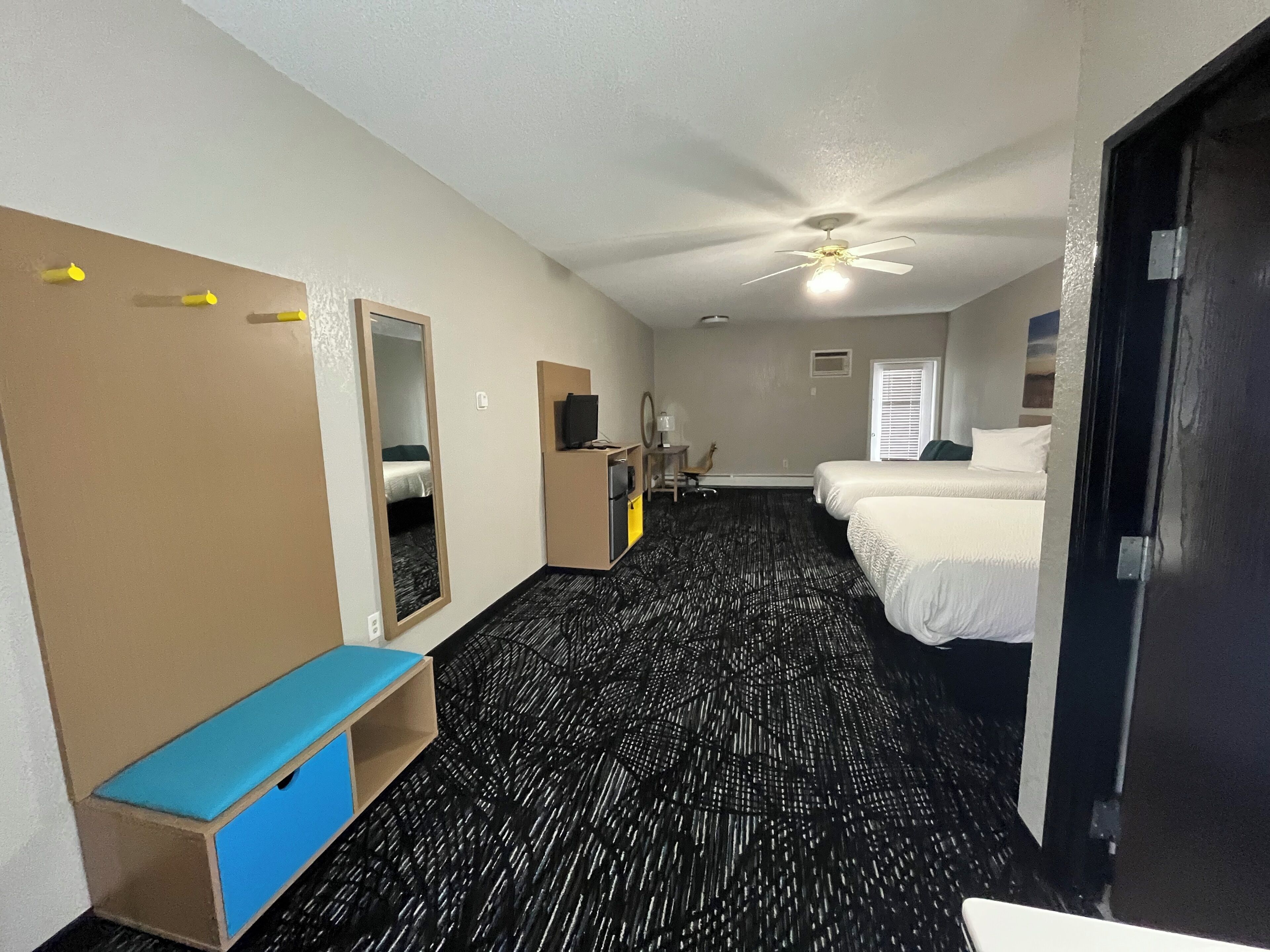 Une chambre d’hôtel équipée d’un lit, d’un bureau, d’une chaise, d’un miroir, d’une armoire et d’un ventilateur de plafond.
