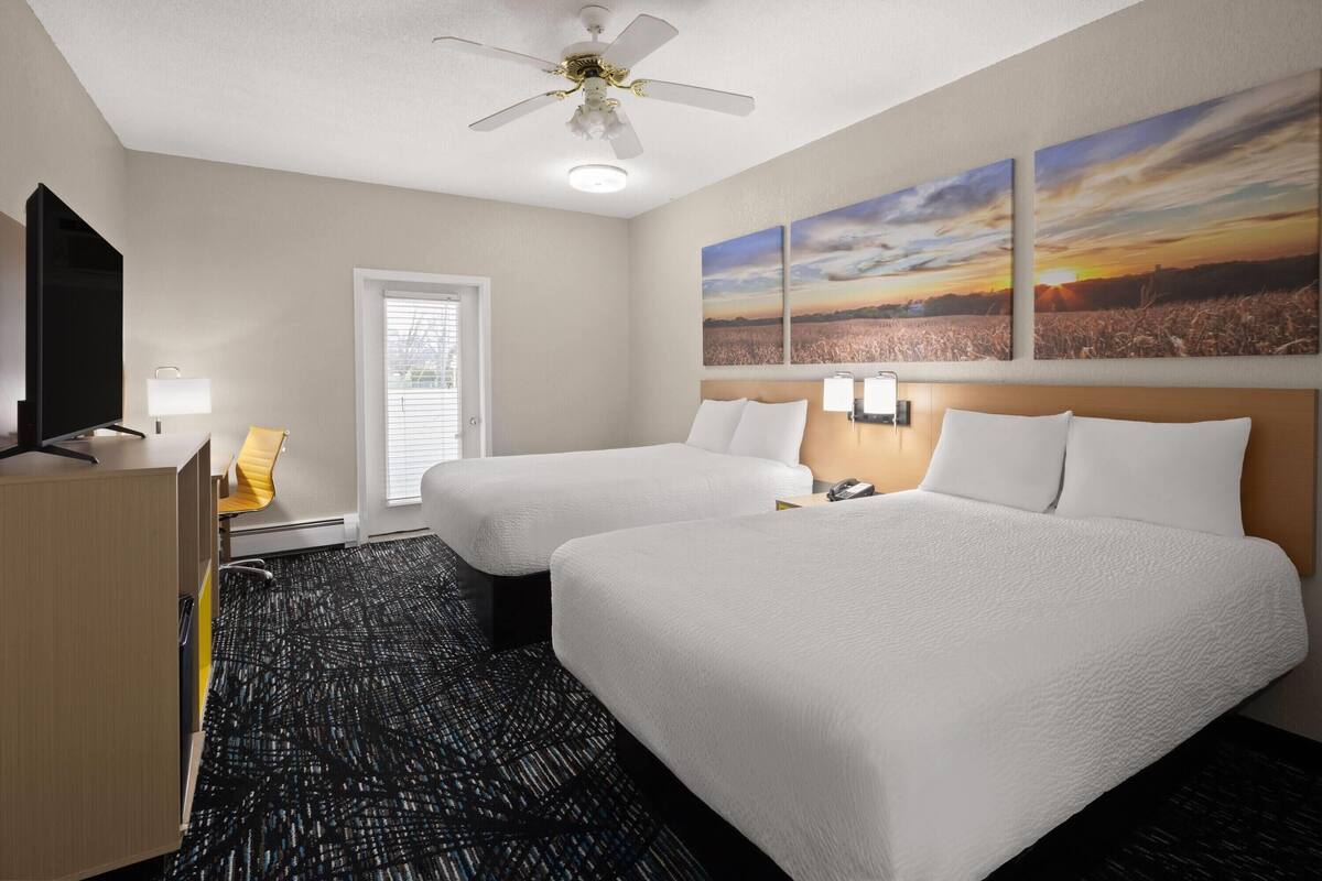 A hotel room with two beds, a desk, a chair, a ceiling fan, and artwork on the wall.