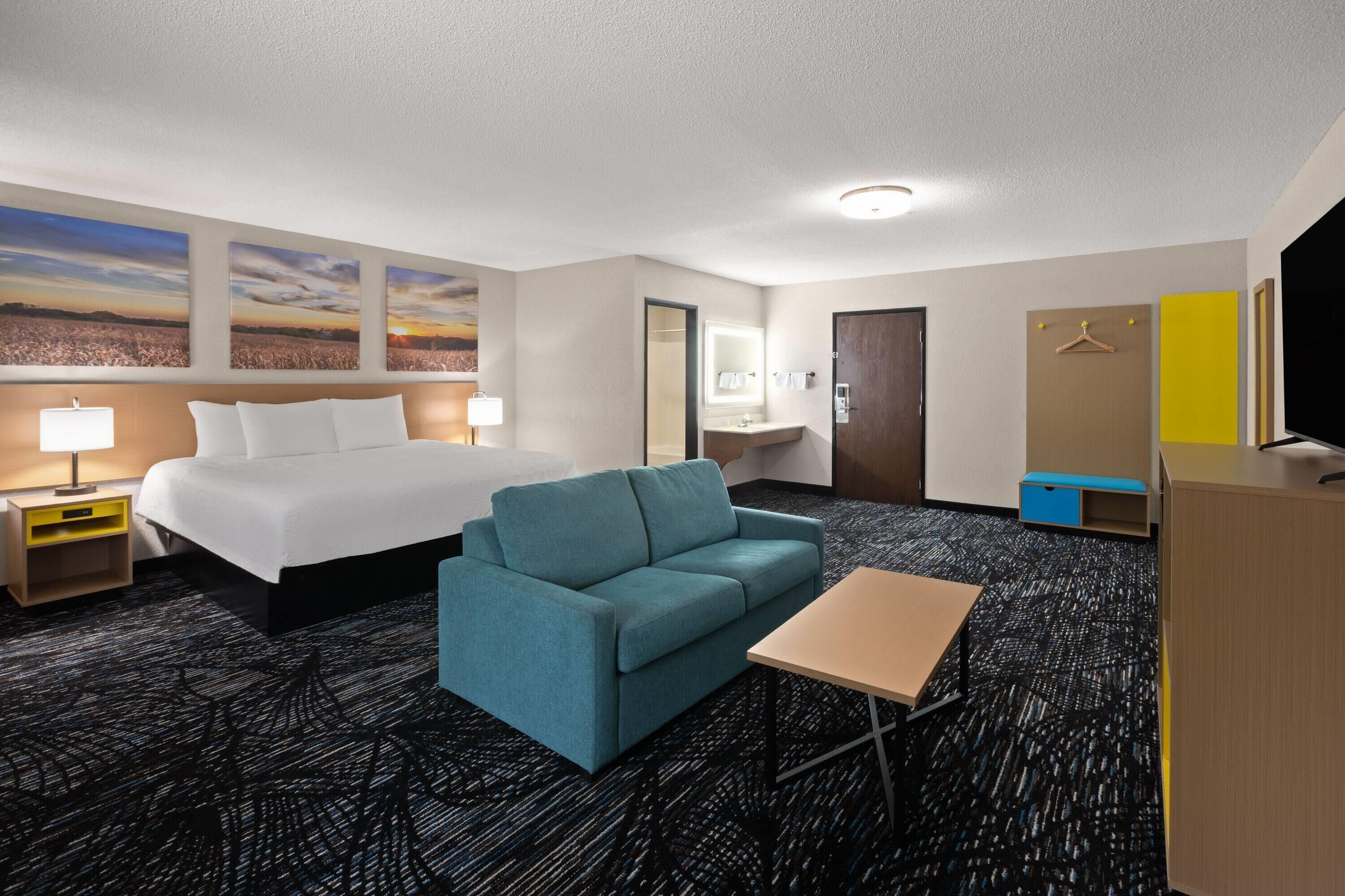 Ein Hotelzimmer mit Bett, Sofa, Schreibtisch, Fernseher, Spiegel und Kunstwerken an der Wand.