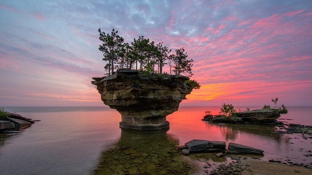 A rock formation with trees on top, surrounded by calm water and a sunset sky.