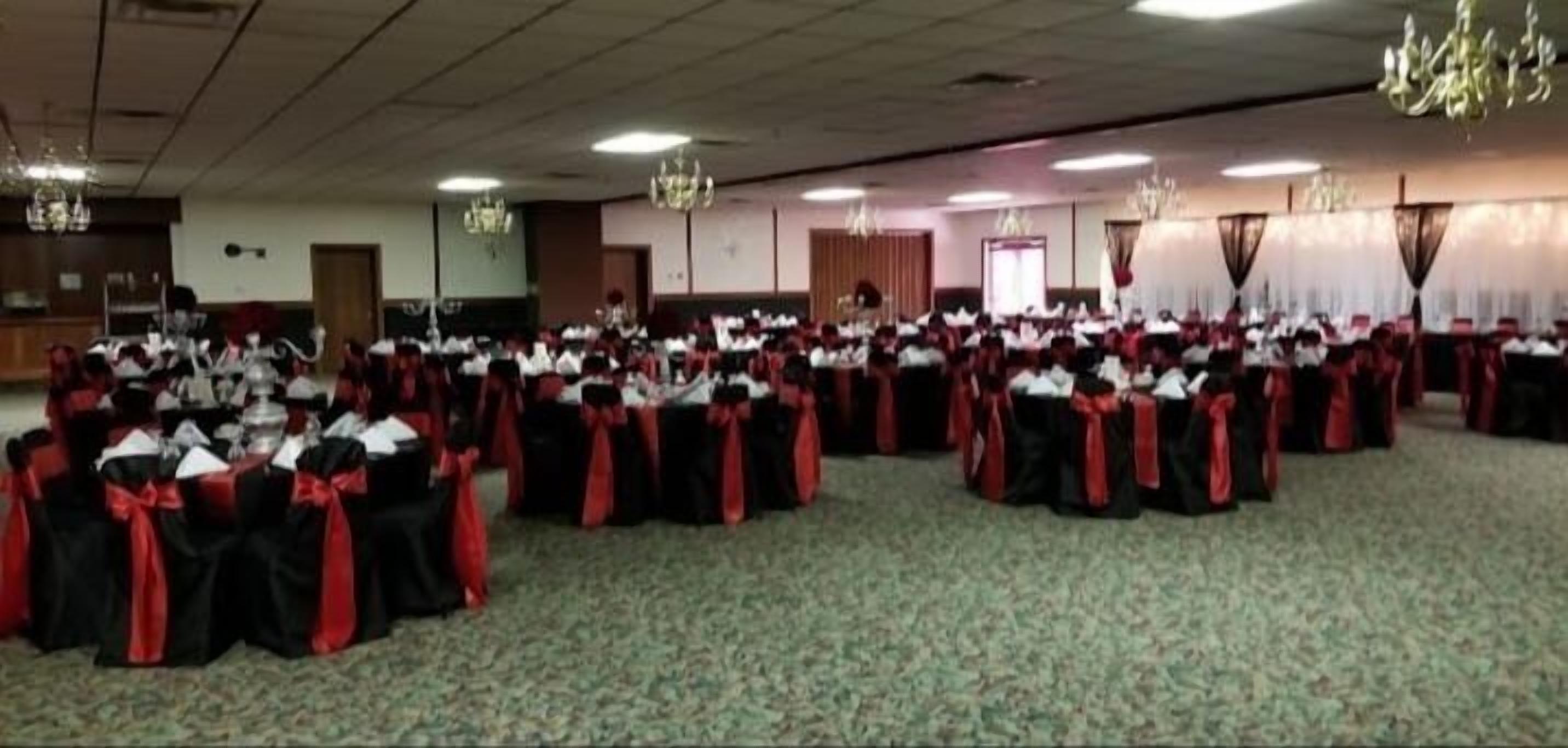 Un salón de banquetes con mesas redondas cubiertas con manteles negros y cintas rojas, dispuestas en filas.