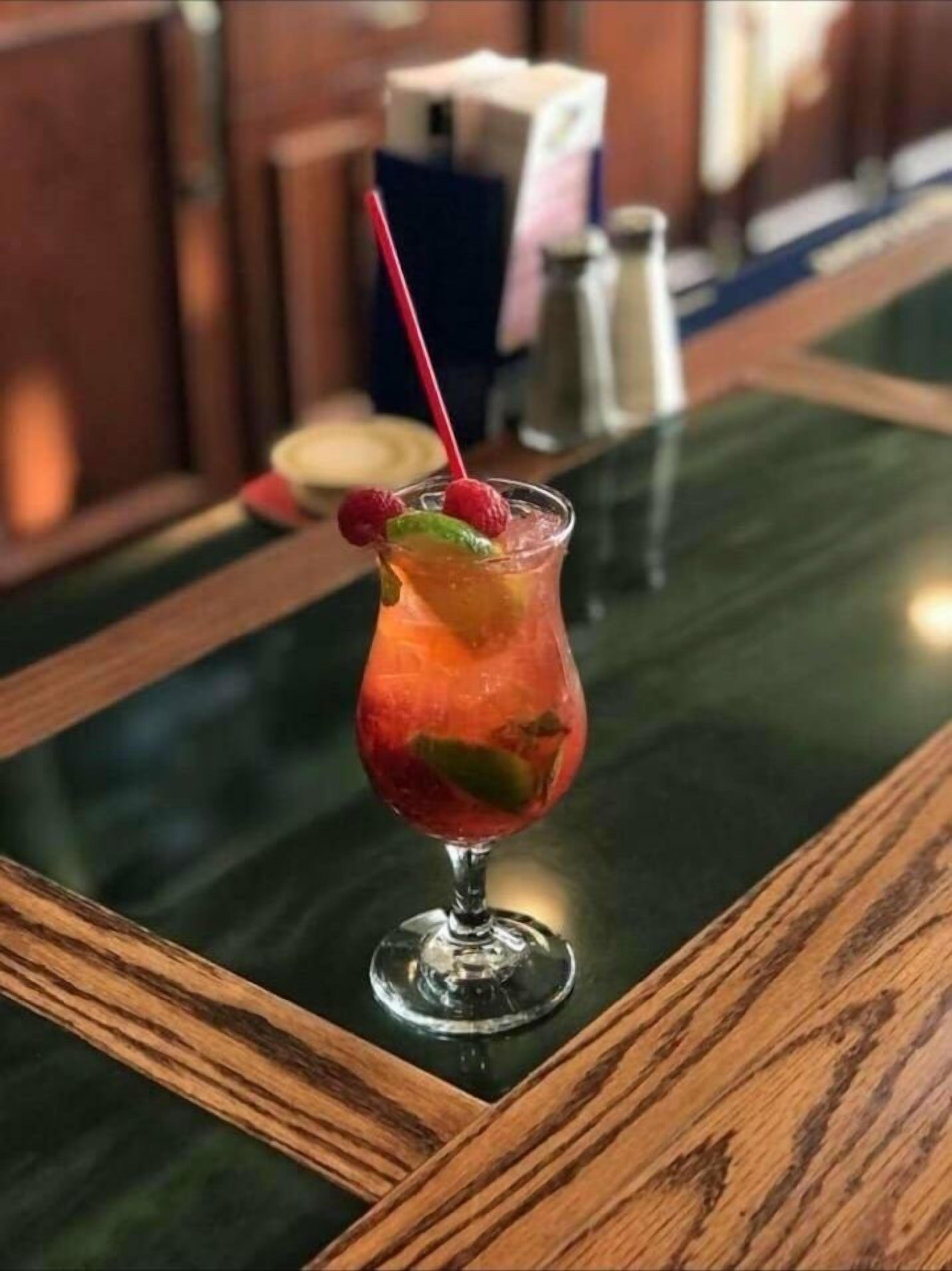Ein Cocktail im Glas mit einem roten Strohhalm und Himbeeren am Rand, auf einem Holztisch mit einer grünen Glasplatte platziert.
