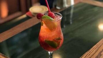 Ein Cocktail im Glas mit einem roten Strohhalm und Himbeeren am Rand, auf einem Holztisch mit einer grünen Glasplatte platziert.