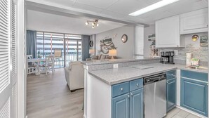 A modern kitchen with blue cabinets, a central island, and a dining area with a view. - Sand Dunes 2451 (Myrtle Beach)