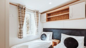 A room with two beds, a window with curtains, a headboard, and a wooden shelf above the beds.