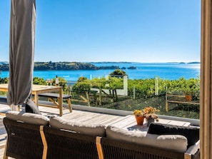 A terrace with wicker furniture, a table with glasses, and a view of the sea and hills. - Island Escape - Sandy Bay Coastal Retreat (Waiheke Island)