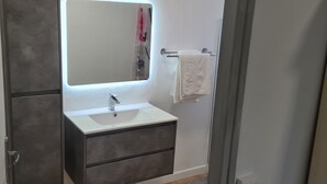 A bathroom with a white sink, dark cabinetry, a mirror with a light, and a towel rack. - Fully furnished and air-conditioned apartment (Les Abymes)