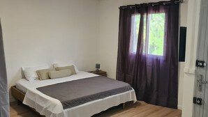 A bedroom with a bed, a bedside table, a window with curtains, and a door. - Fully furnished and air-conditioned apartment (Les Abymes)