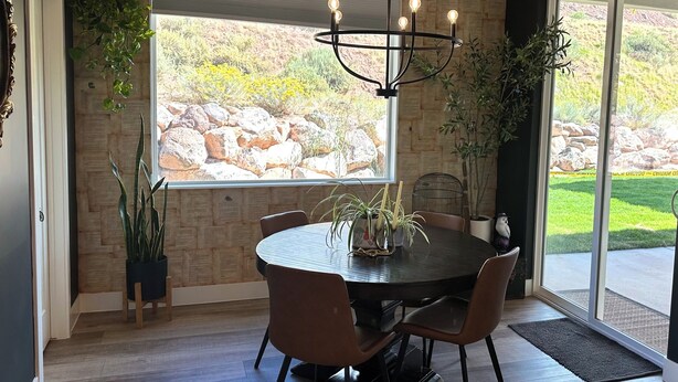 A dining area with a round table, chairs, a chandelier, and large windows with a view of rocks and greenery.
