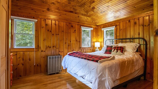 A wooden-paneled bedroom with a bed, two windows, a heater, and a lamp.