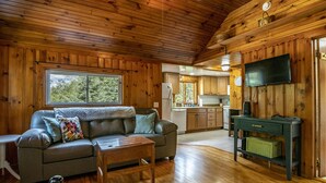 A cozy cabin interior with a wooden ceiling, a sofa, a coffee table, a television, and a kitchen area.