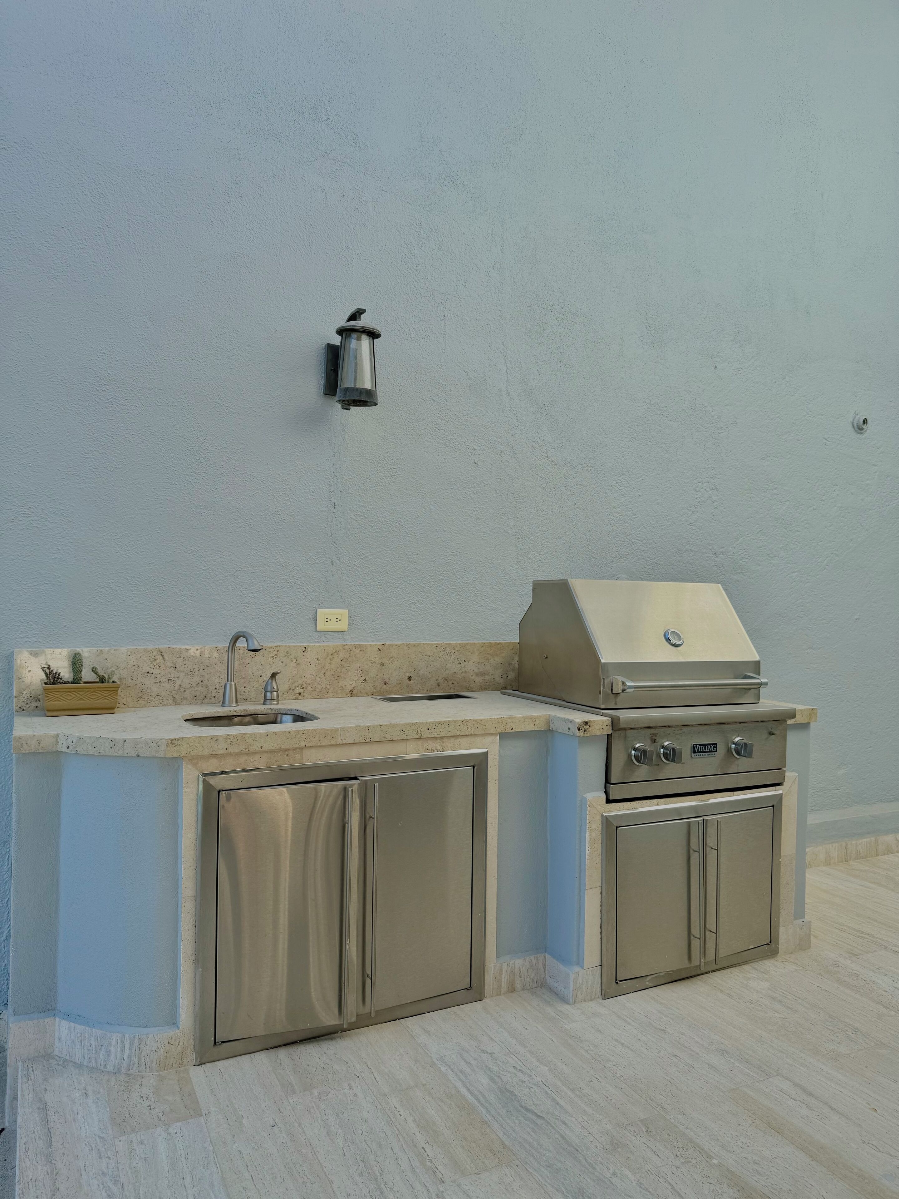 An outdoor kitchen with a sink, a built-in grill, and a wall-mounted light fixture.