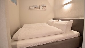 Una cama pequeña y ordenada con ropa blanca en una habitación compacta, con techo inclinado y ventana pequeña.