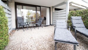Un patio con dos sillas, una mesa y una puerta de vidrio que da a una habitación, con un seto a la derecha.