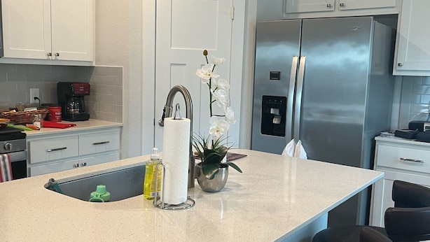 Une cuisine moderne avec des armoires blanches, un réfrigérateur en acier inoxydable, un îlot central intégré avec des tabourets, et une petite orchidée en pot.
