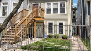 Un immeuble résidentiel de deux étages, doté d’un escalier en bois menant à la porte d’entrée, d’un petit jardin devant avec une clôture en métal et d’un arbre aux fleurs roses.