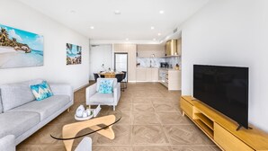Een moderne woonkamer met een grijze bank, een salontafel en een televisie op een houten meubel. In de achtergrond bevindt zich een keuken met witte kasten en een eethoek met een tafel en stoelen.