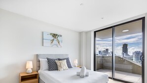 Een moderne slaapkamer met een groot bed, twee nachtkastjes met lampen, uitzicht op de stad door een glazen deur en een schilderij aan de muur.