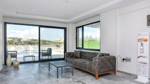 A modern living room with a grey sofa, a small table, and a view of a pool and outdoor seating area through large windows. - Beyaz Konak Otel (Ayvalik)