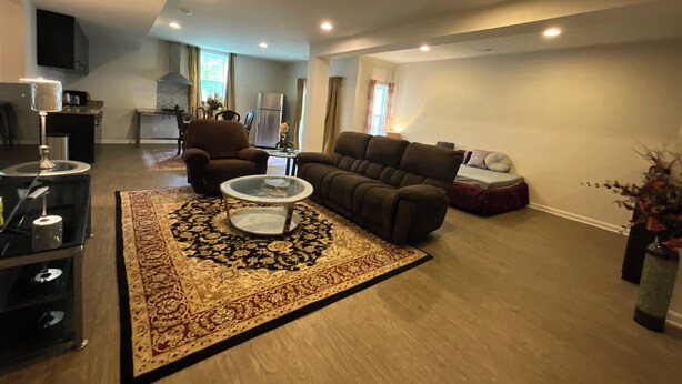 Un salon spacieux comprenant un canapé, un fauteuil, une table basse et un tapis. Au fond, on aperçoit un coin cuisine avec un réfrigérateur et une table à manger.