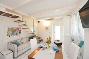 Un salon moderne avec un canapé, une table basse et un coin repas avec une table dressée pour deux personnes. Un escalier avec une rampe en verre mène à l’étage supérieur.